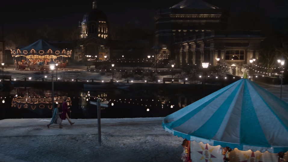 Calah Lane and Timothée Chalamet run along the path in Verulamium Park in the Wonka movie. There is a fairground stall in the foreground and CGI buildings on the other side of the lake.