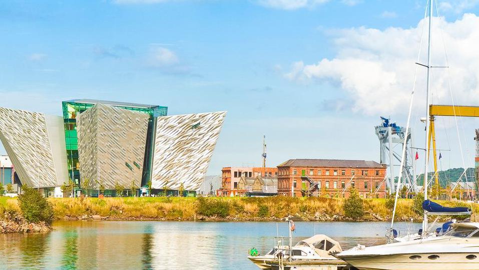 Titanic Quarter in Belfast. Sea in the foreground. To the right, a partially visible view of a large, yellow crane, and left across the frame, a brick building, the Titanic Belfast building and industrial buildings.