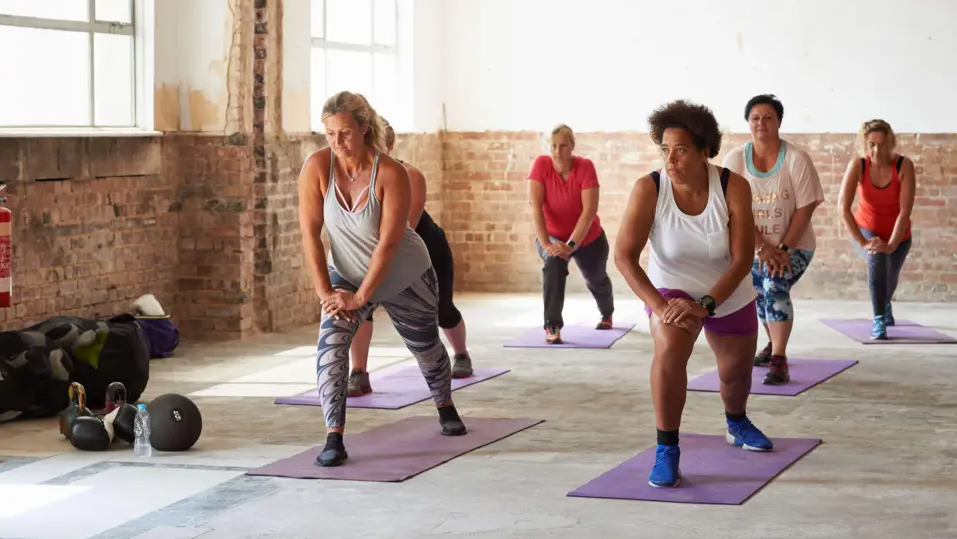 Six women do exercises in a warehouse-style gym environment, with exposed brick walls and concrete floors. They are wearing a mix of vest tops, T'shirts, leggings and trainers, and are all standing in a lunge position with their hands on the top of their right leg on purple mats.