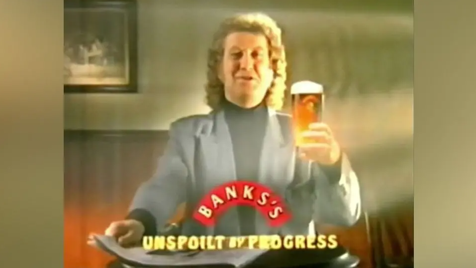 Noddy Holder is pictured holding a pint of Banks's beer and reading a paper in a TV advert. He has curly hair and is wearing a black jumper and grey jacket.
