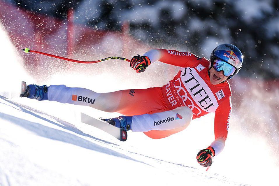 An alpine skier in a red racing suit carves through fresh snow at high speed on a downhill slope