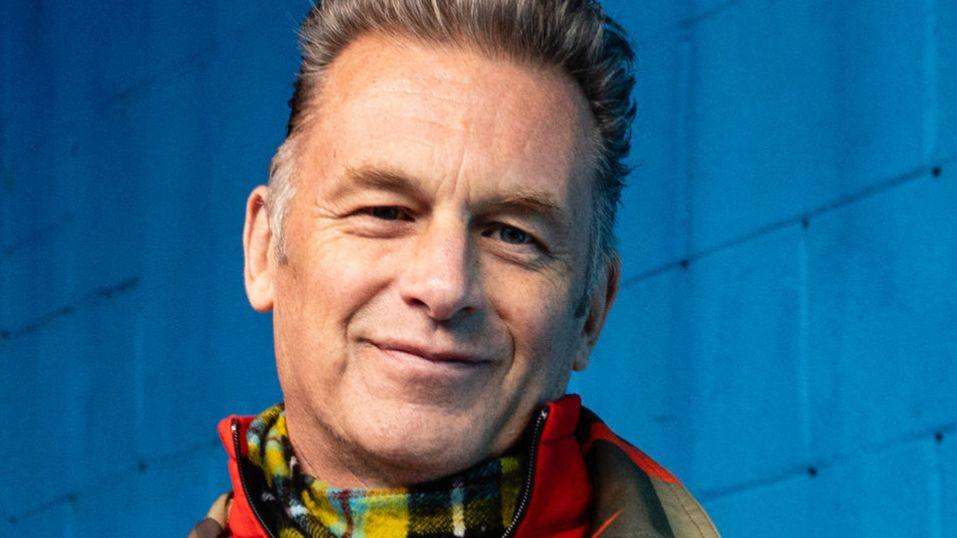 Chris Packham headshot wearing a chequered scarf and a red jacket. Behind him is a blue wall