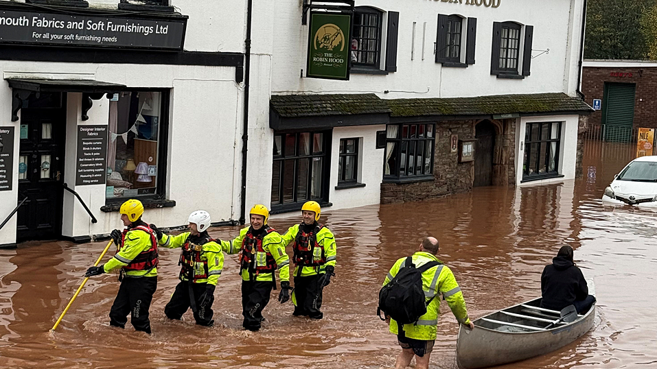 Four rescuers in hi-vis wade through flooded street as a man in a hi-vis jacket pushes a person in a canoe. There is a pub on the left of the image and a shop. They are both white buildings.