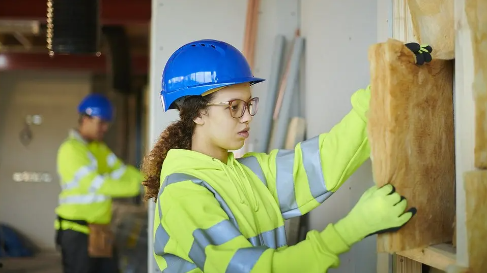 A woman in a hard hat and fluorescent jacket handles insulation inside a building. A man is in the background in a similar jacket and hat.