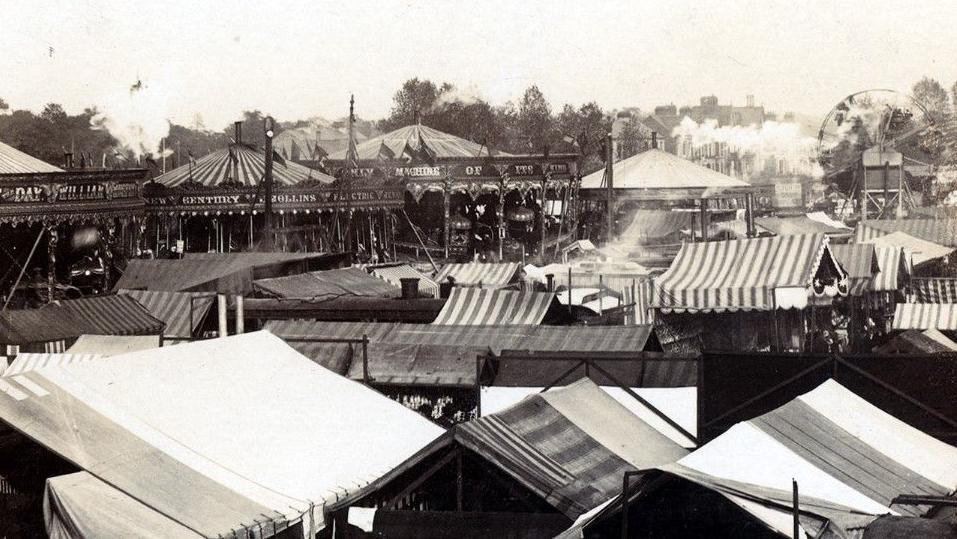 A black and white picture of a fair with tents and waltzers in the foreground and a big wheel in the background.