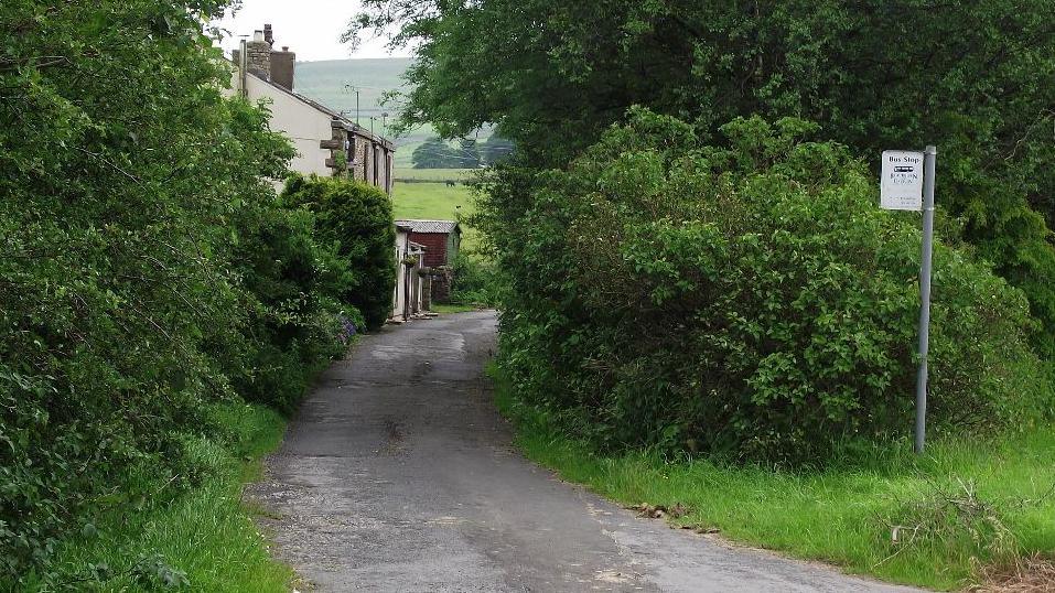 Bus stop by an empty lane in Tockholes, which is lined by green shrubs, and an old village house in the background, with green Pennine hills in the backdrop.