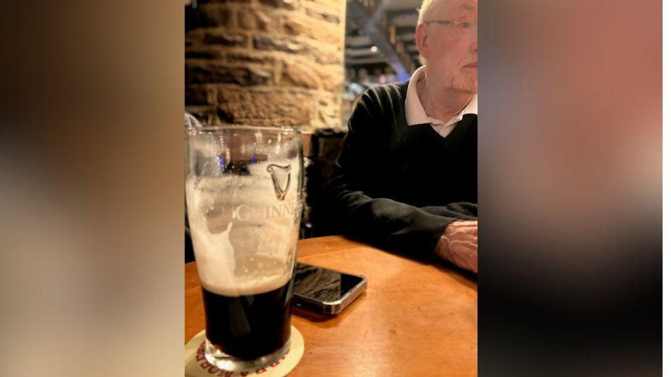 A close-up of a pint of Guinness that is only one third full, in front of a mobile phone. To the right in the background an elderly gentleman sits.