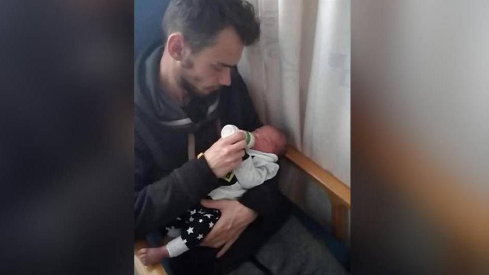A man with brown hair holding a baby in his left arm. With his right hand, he is feeding the child with a bottle of milk. They are sitting in a chair with a curtain visible on the left of the pair of them.