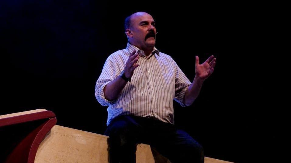 Paul Garrett, on stage, with a moustache and brown hair wearing a white striped shirt and a watch