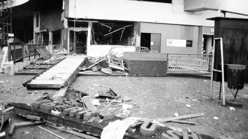 The pub is seen after the blast. Windows are blown out, debris is strewn across the floor, the pub sign is on the ground in the black and white image.