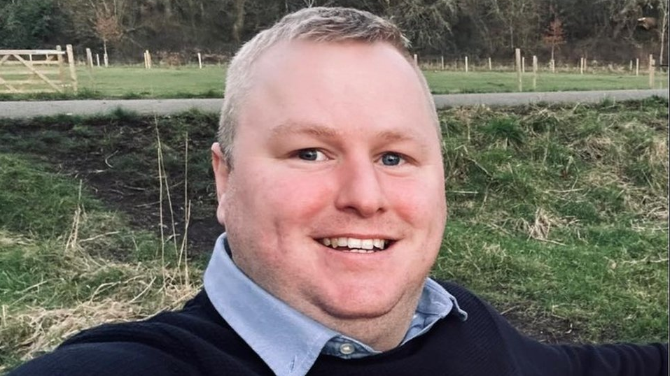 Darren Brown, with short brown hair, a white shirt and navy jumper 