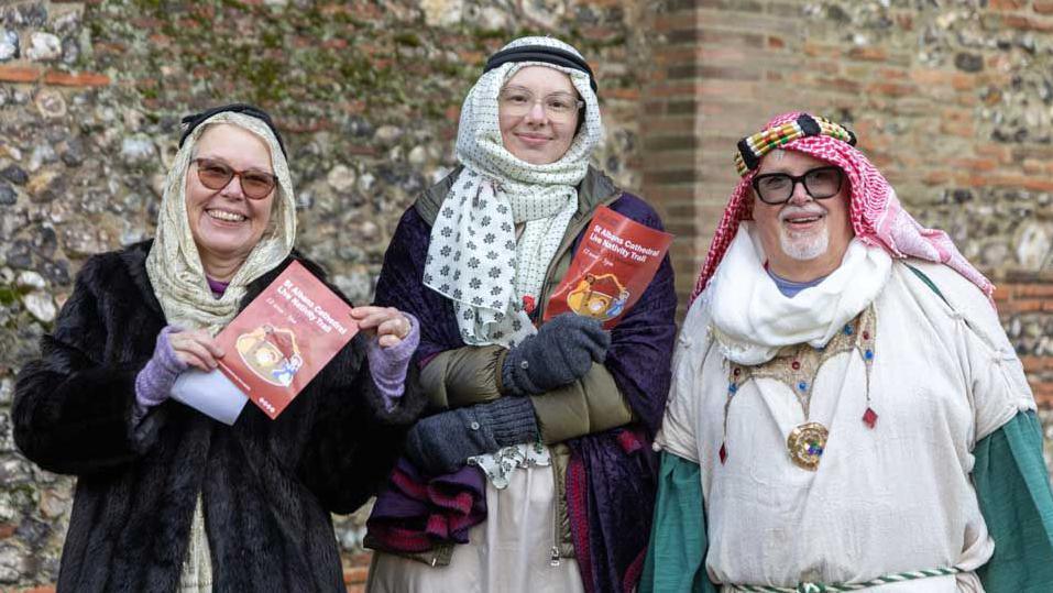 The three wise people – a woman on the left in a black coast and beige headscarf holds a leaflet for the nativity trail, as does a woman in the middle wearing a purple shawl and a beige headdress. A man on the right is in a white tunic over the green shirt with a red and white head-dress