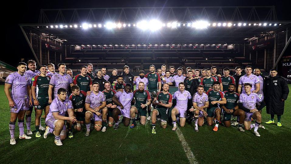 Gloucester and Leicester players stand and kneel together on the pitch with Tommy Reffell in the centre holding the Slater Cup