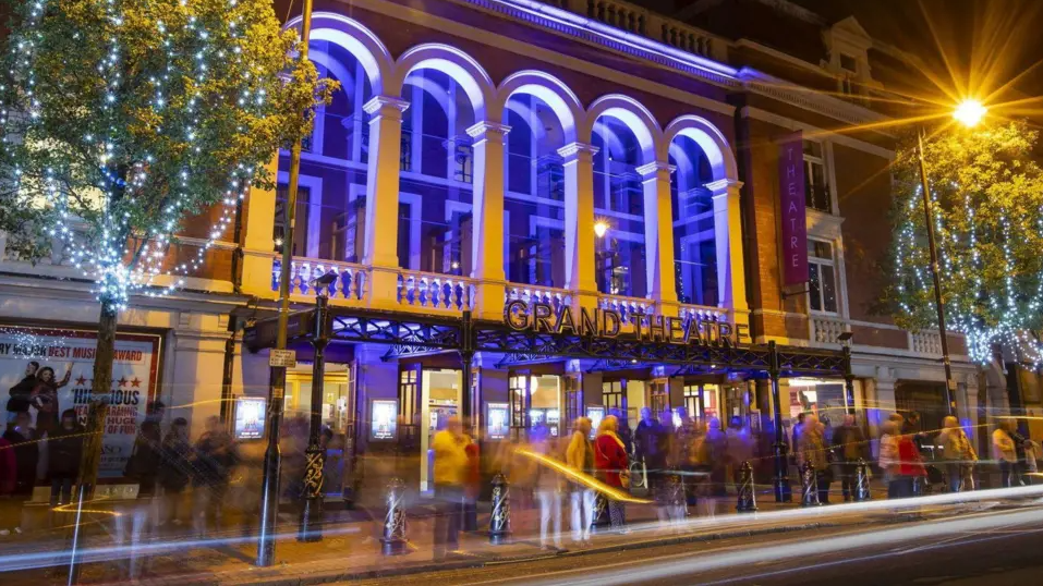 The theatre is lit up at night with people walking by outside. It has a purple hue from the lighting  with two trees either side with lights on.