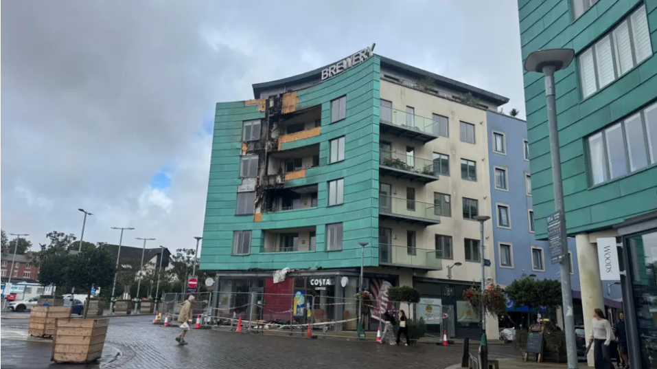 A photo of the building in the aftermath of the fire in the town square. The block of flats is partially blackened by the smoke. The building has been cordoned off and there are orange and white cones in front of it. Some people are walking nearby. It is cloudy.