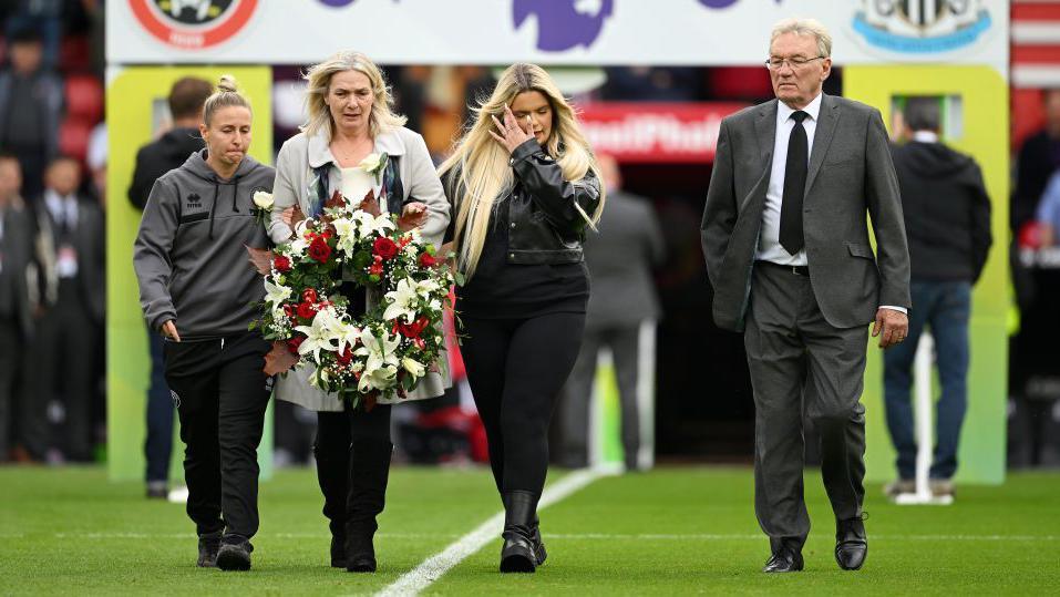 Three women and a man walk side-by-side into the centre circle of a football pitch. One of the women, wearing black trousers and a grey overcoat, carries a large wreath of white and red flowers at waist level. Next to her a younger woman in a black leather jacket appears to wipe a tear from her eye, while the man, in a grey suit with white tie and black shirt, looks forward.