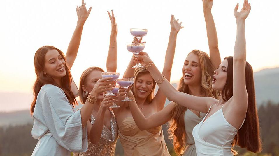 Young women enjoying a bachelorette party, wearing elegant dresses drinking a lavender drink outdoors at sunset, raising hands.