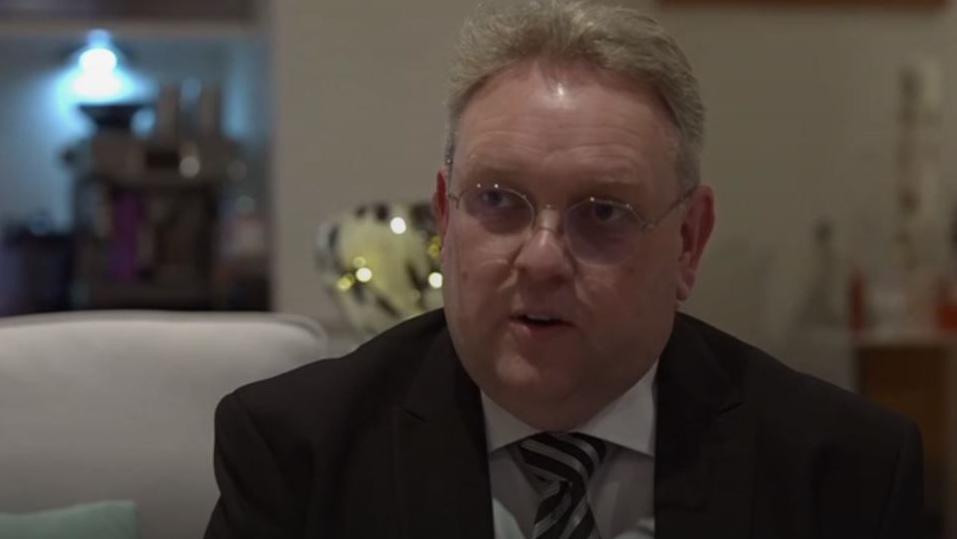 Matthew Ineson sits on a sofa in silver-rimmed spectacles, a black suit, white shirt and grey and black striped tie