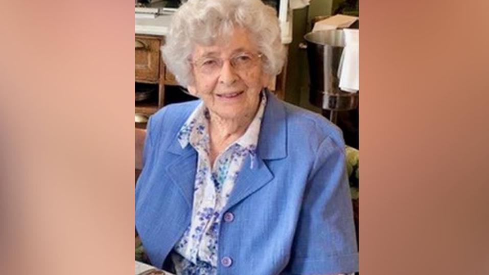 Margaret Packham is sitting in a restaurant smiling at the camera. She is wearing a bright blue jacket over a purple and blue blouse and a pair of glasses.