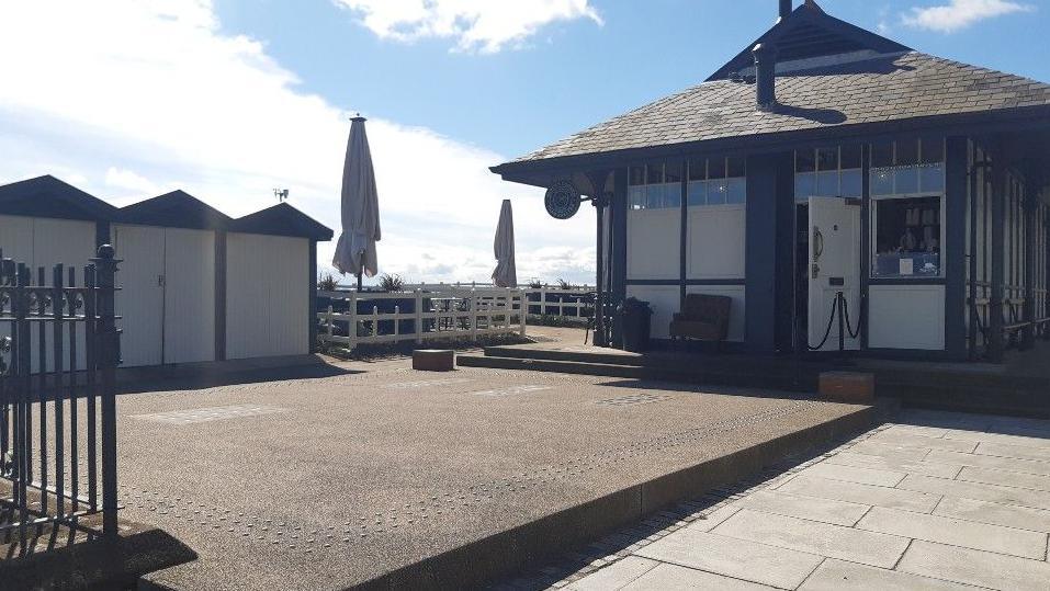 The patio and side entrance of the former tram shelter, showing three black beach huts with white painted doors.