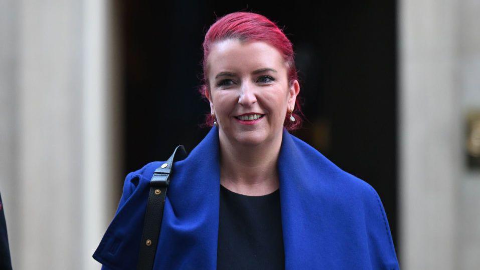 A woman with long red-dyed hair smiles. She has a blue coat on with a black top underneath. She has a black leather bag over her left shoulder. She has to small gold earrings