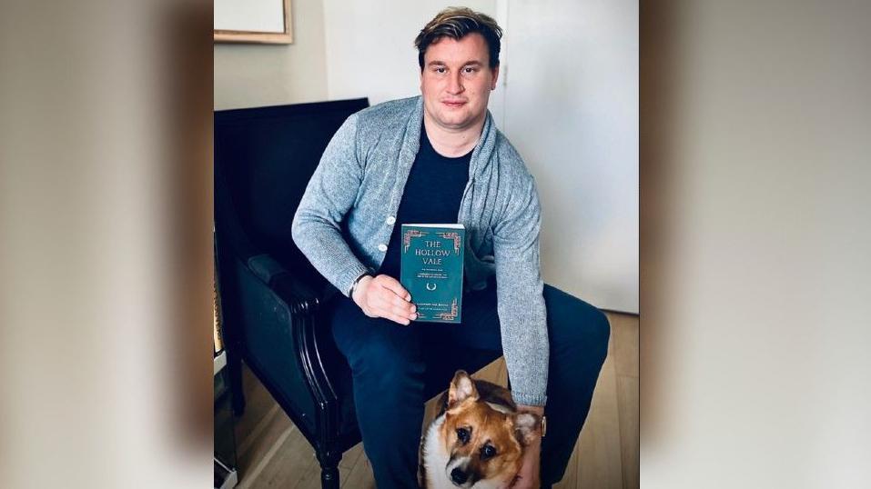 Alexander holding a copy of his book in one hand and patting his dog - a corgi - with his other hand. Alexander is wearing a blue shirt over a dark top and jeans.