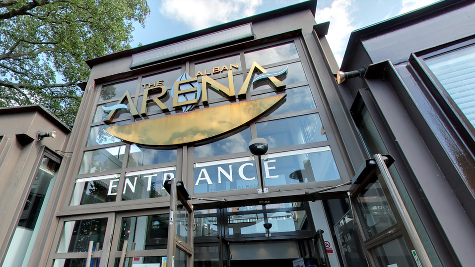 The entrance to the Alban Arena in St Albans is made of steel and glass with a large gold sign depicting the venue's name and the word "entrance written in white.