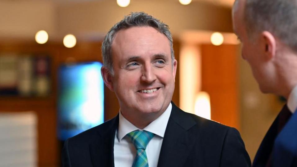 Alex Cole-Hamilton smiling at a man who is out of focus. He has grey hair and is wearing a black suit, white shirt and green tartan tie.