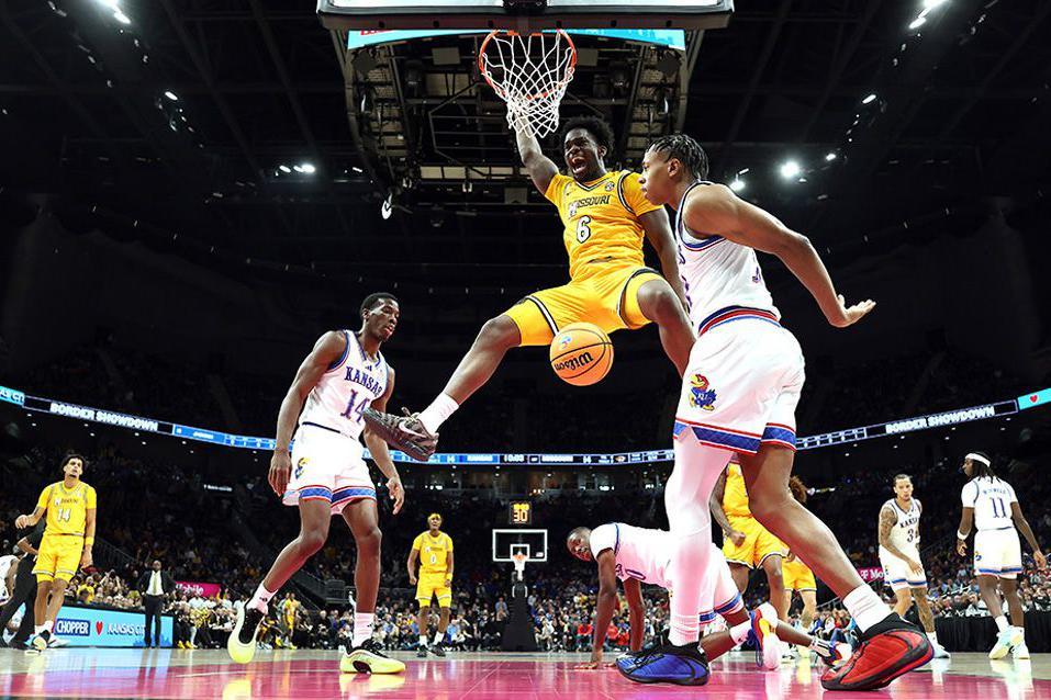 Annor Boateng of the Missouri Tigers in a yellow uniform hangs from the basketball rim with a look of excitement on his face after completing a powerful dunk