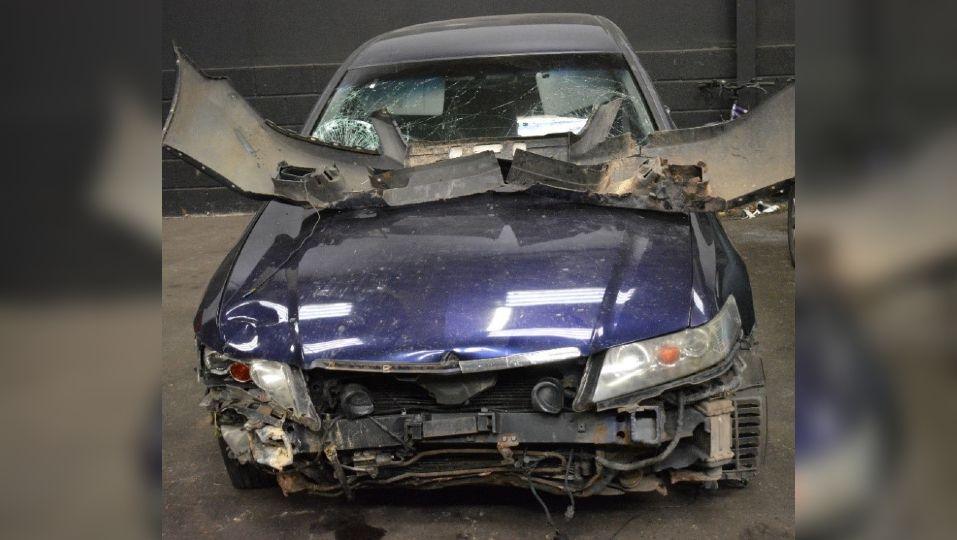 The blue Honda Accord following the incident. The front of the car is smashed and crumpled from the impact. The bumper has been torn off and is resting next to the windscreen, which has been smashed in multiple places.