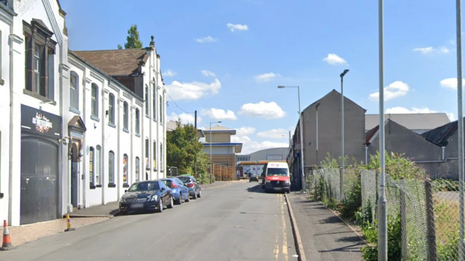 There are cars parked on with side of the road in Frederick Street with buildings on one side and grassland bordered by a fence on the other.