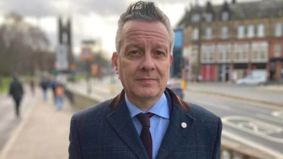 Nick Kemp, with short grey hair is standing on a high street. He is wearing a blue suit and is looking at the camera, expressionless.