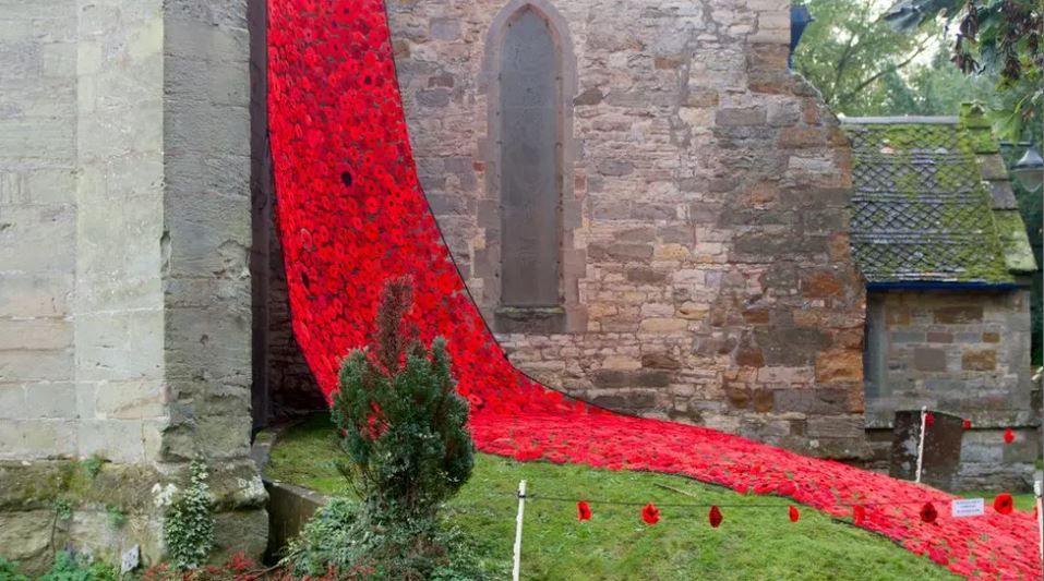 Hundreds of red knitted poppies hang down the side of a grey stone and brick building with a narrow archway in the middle. Some poppies are also covering the grass.