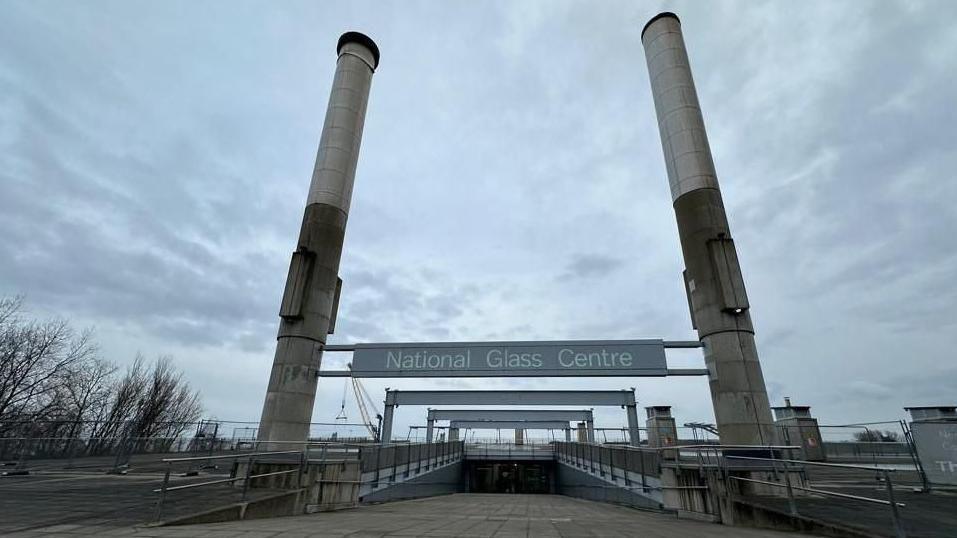The entrance to the National Glass Centre. The concrete path leads down a ramp. Meanwhile, two concrete pillars border either side. A grey sign reads: "National Glass Centre".