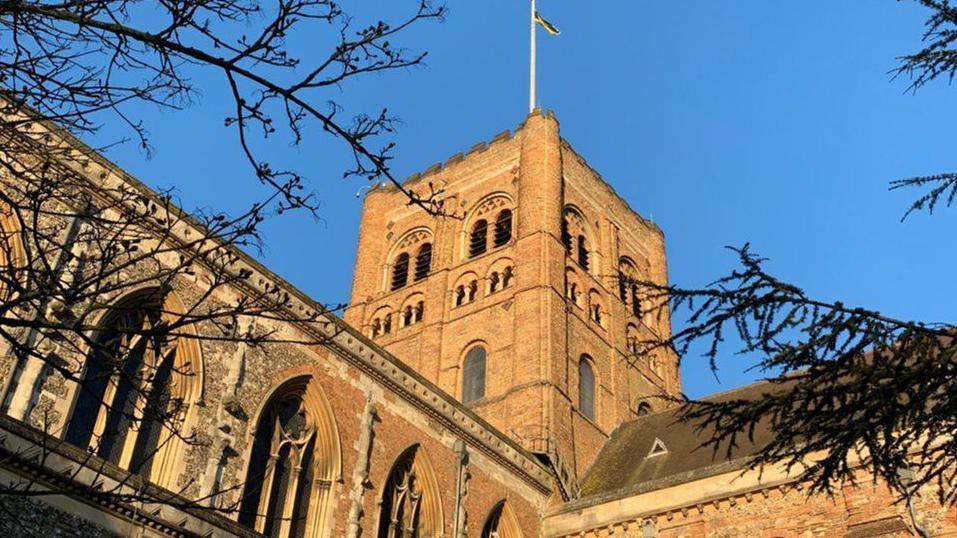 The brown brick of St Albans Cathedral's Norman tower is seen against a bright blue sky. There are fir tree branches in the foreground.