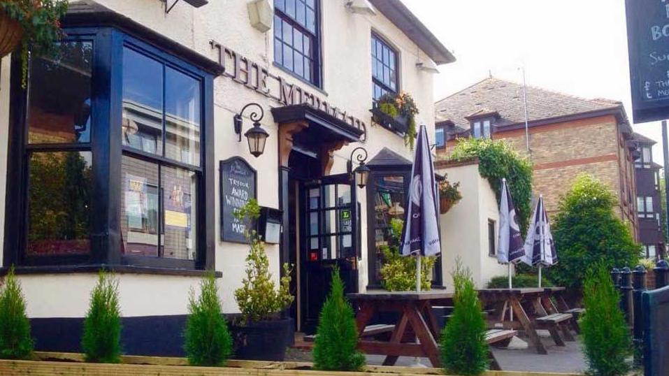 The outside front view of The Mermaid pub in St Albans, Hertfordshire with some wooden tables and umbrella's outside