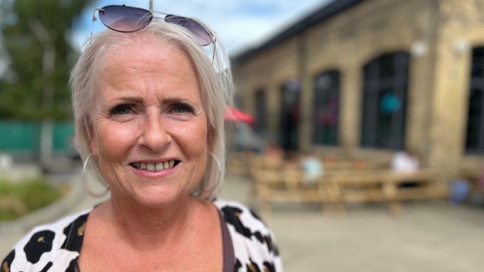 Karen Perks is smiling into the camera. She has fair hair that is just above shoulder length and has sunglasses perched on her head. She is wearing a black and white patterned top. A brick building - a pub - can be seen blurred in the background