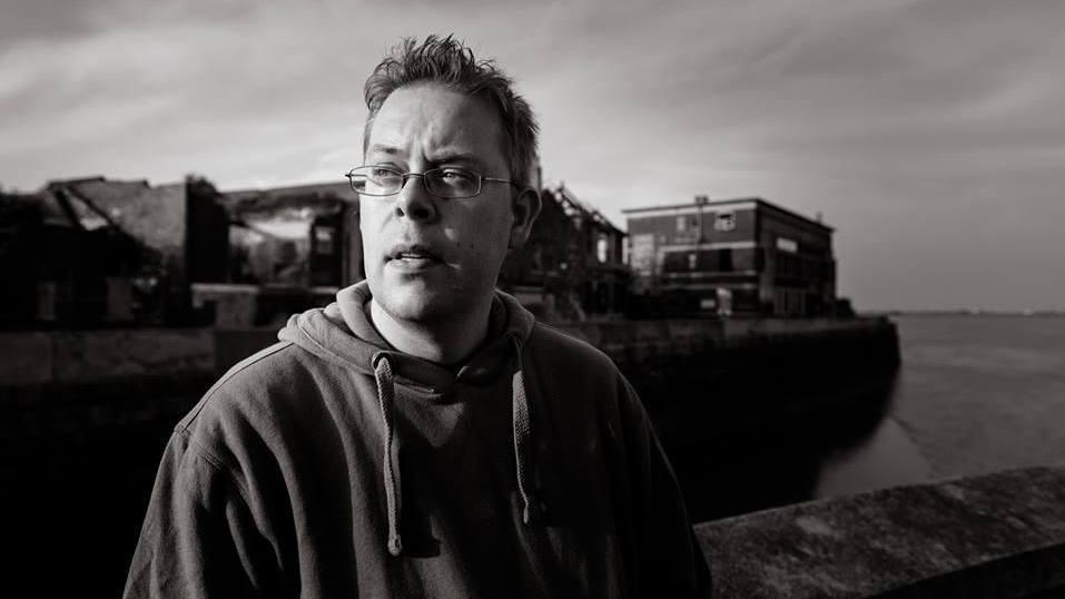 Nick Quantrill. A moody black and white photograph taken outside with a boat in the background.  He wears a hoodie and glasses.