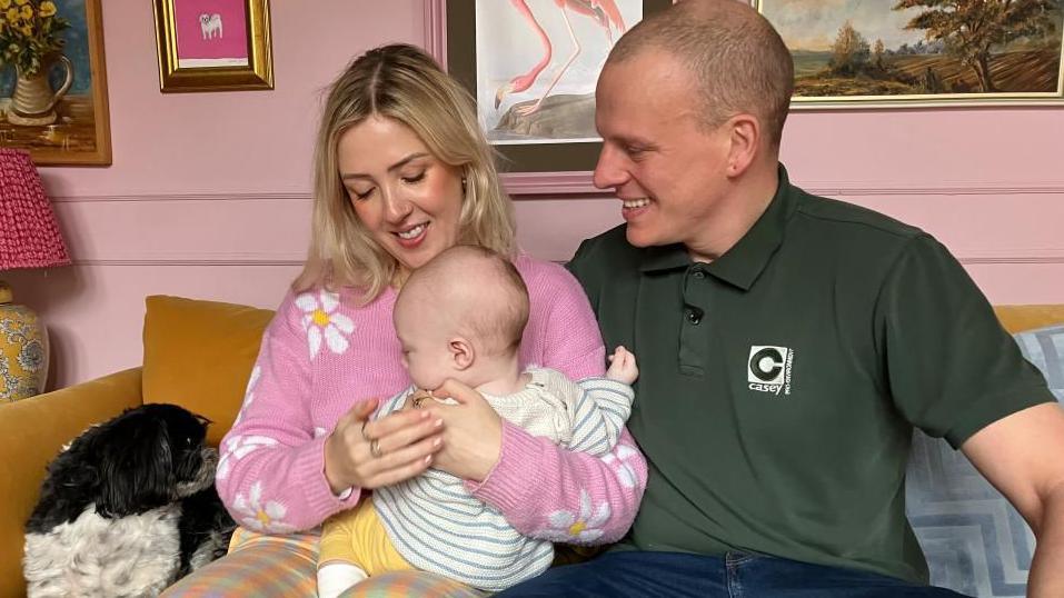 Jessica and Rick Sowerby sit on a sofa with their baby son on Mrs Sowerby's knee. Mrs Sowerby has bobbed blonde hair and is wearing a pink jumper with a daisy pattern. Mr Sowerby wears a green polo shirt. Both are smiling and looking at their son, who wears a grey and white striped jumper, yellow trousers and white socks. The couple's dog Merla can be seen on the sofa beside them.