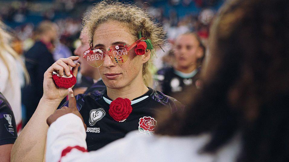 Abby Dow wears some heart shaped sunglasses with her name written on them and a crochet red rose on her rugby top.
