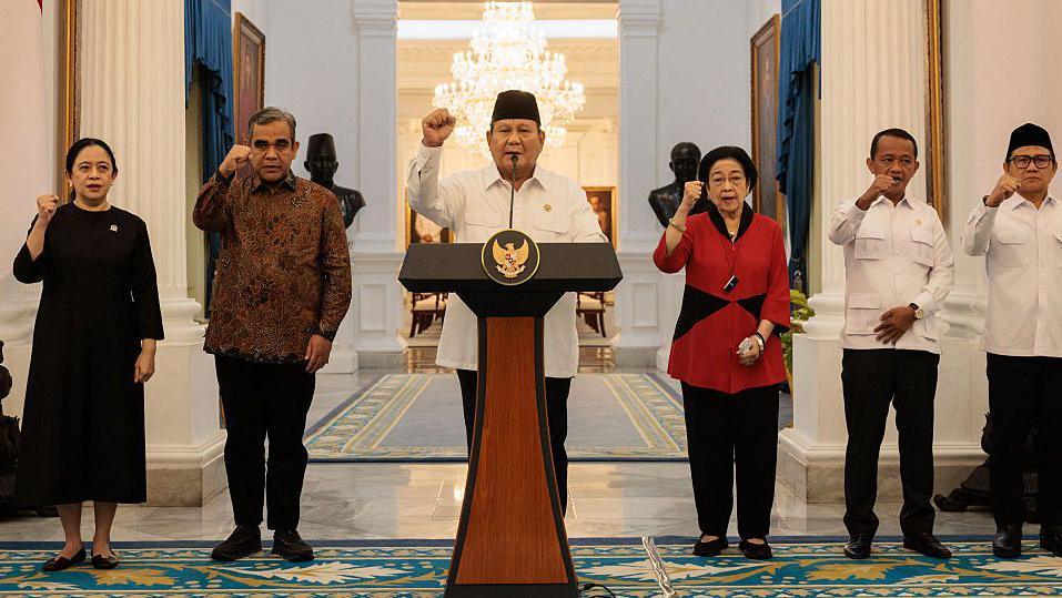 President Prabowo Subianto flanked by political party leaders inside Jakarta's presidential palace. Prabowo is speaking at a lectern, with his fist raised - a gesture copied by the other political leaders in the photo