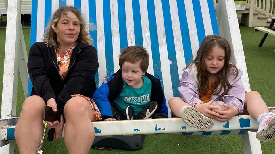 A woman sits on a giant, blue and white deck chair next to a young boy and young girl.