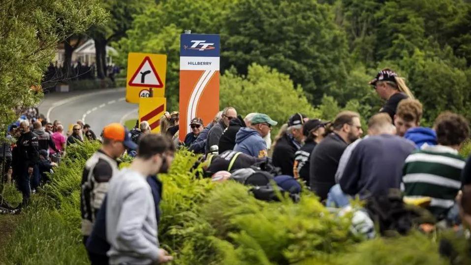 Spectators lining the hedge at Hillberry on the TT course. A sign for the milestone can be seen alongside other road signs, as the road sweeps up to the right.