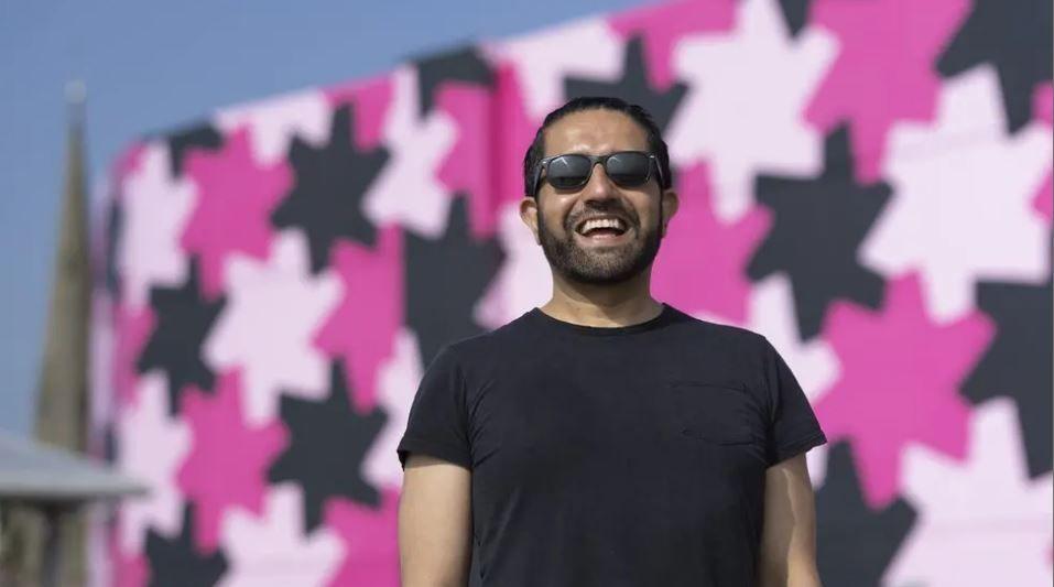 A man wearing black sunglasses and a black t-shirt stood smiling in front of the backdrop of a building featuring baby pink, hot pink and black star shapes.