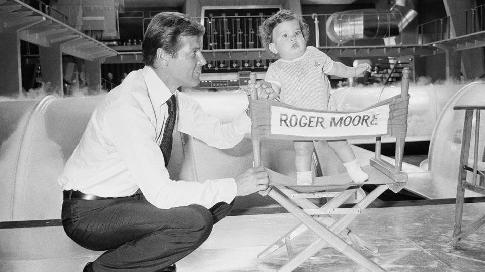 In this black and white image, Sir Roger is seen on the film set of the Bond film, The Man With The Golden Gun. He is crouching down next to a chair which has his name written on the back. His young son Christian is standing on the chair and Sir Roger is holding his hand. In the background, you can see pipes and tunnels.