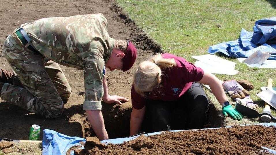 Military personnel in uniform and member of the dig team reaching into an excavated hole in the ground with spades and other items scattered around the site