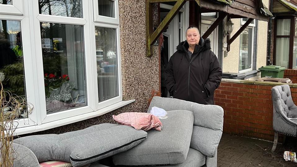 Dawn Royds , who is wearing a black coat, with the furniture in her front garden following the floods on Templeton Road