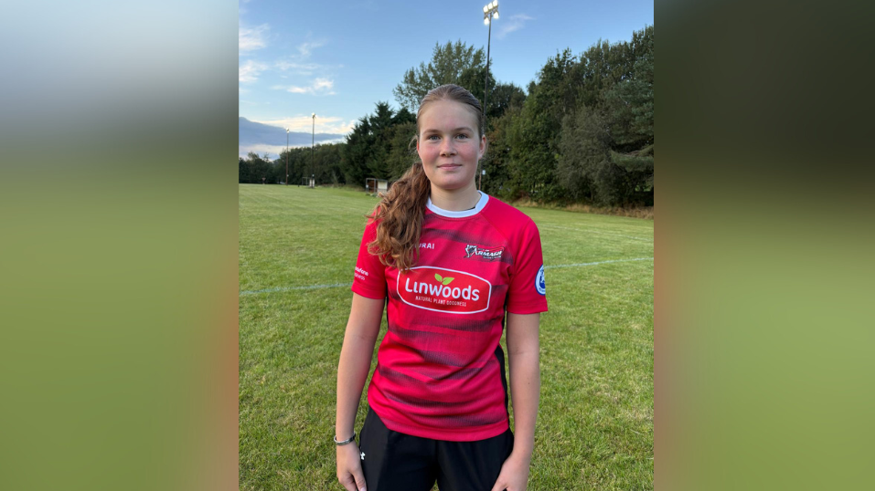 Eva standing on the pitch, she's wearing a red rugby top that says Armagh on it alongside sponsors. Her red/brown hair is tied up in a ponytail and she is smiling. 
