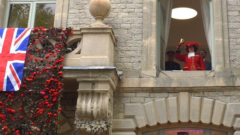 A town crier standing at the upstairs window of a grand stone building. She is wearing red ceremonial robes. Over the entrance to the building is a blanket of poppies overlaid with a union flag