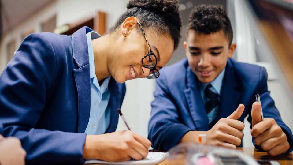 A girl in the foreground is writing on a pad and smiling while looking at the pad. She wears black-rimmed glasses with tortoiseshell sides and a royal blue school uniform blazer with a light blue shirt. A boy next to her smiles at what she is writing. He wears the same uniform but has a dark blue tie.